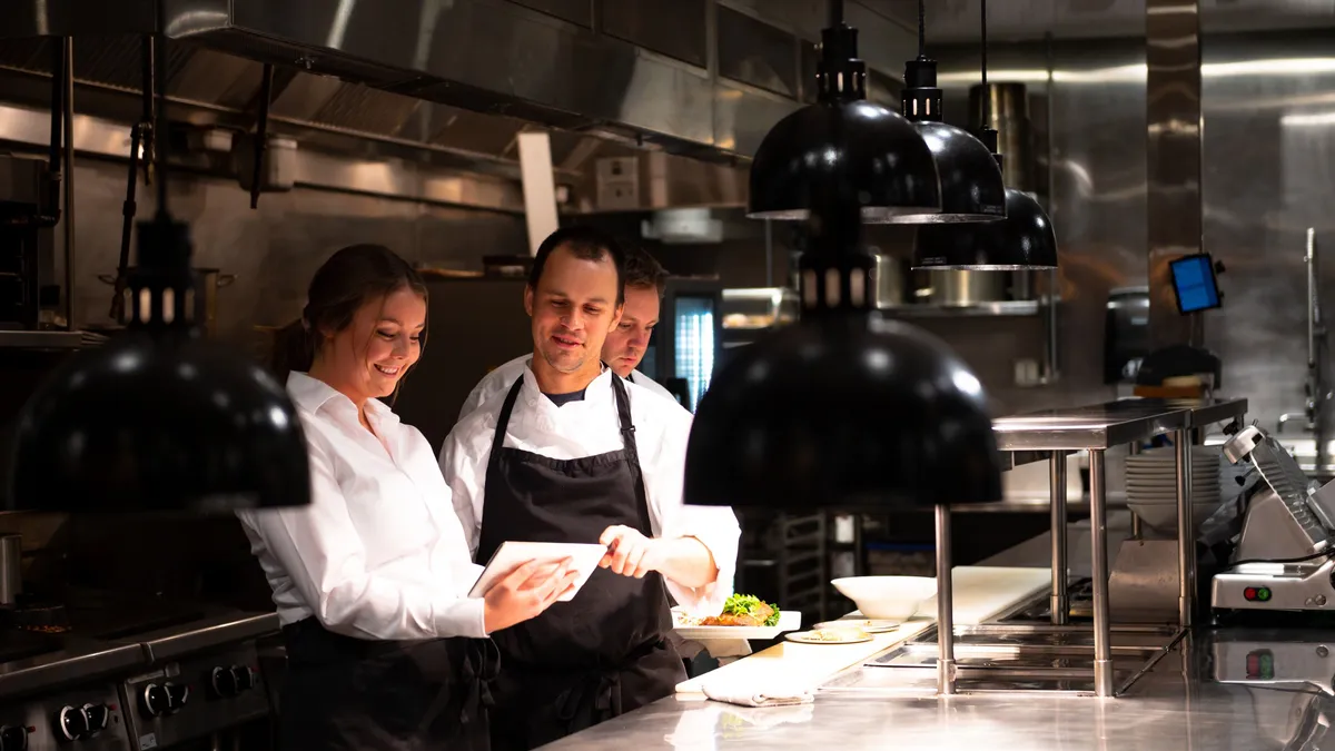 Three chefs in the kitchen. Tow are looking at a tablet and smiling.