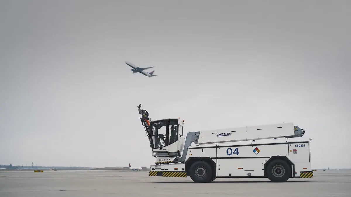 A deicer vehicle parked  on an empty airport runway and an airplane flying overhead.