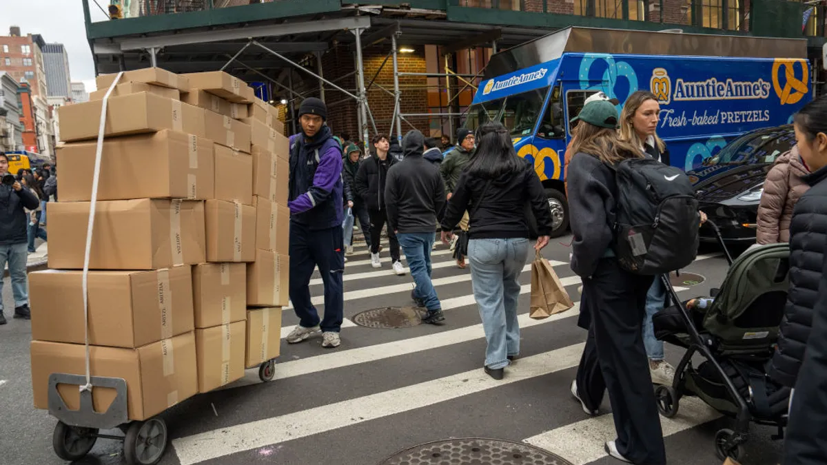 A person pushing a cart stacked with boxes navigates around people on a busy crosswalk.