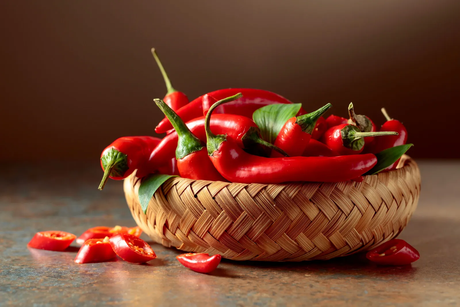 A photogrpah of red chili pepers with green tops in a whicker a basket on a granite table.