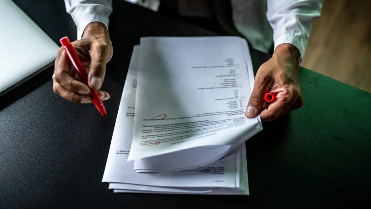 Hands are shown holding a bunch of papers. A red pen is in one hand and there are red marks on the top paper.