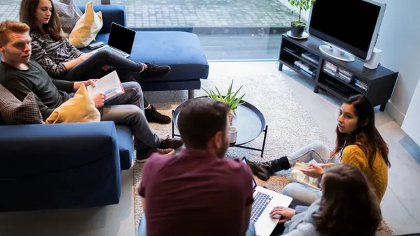 People sit on couches in a living room, holding books and laptops.