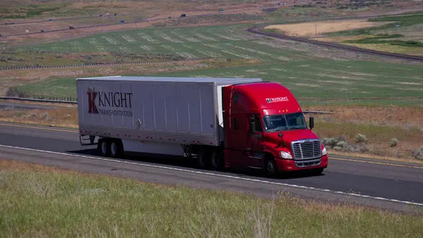 A Knight Transportation Freightliner Cascadia tractor-trailer on a highway with farming areas in the background.