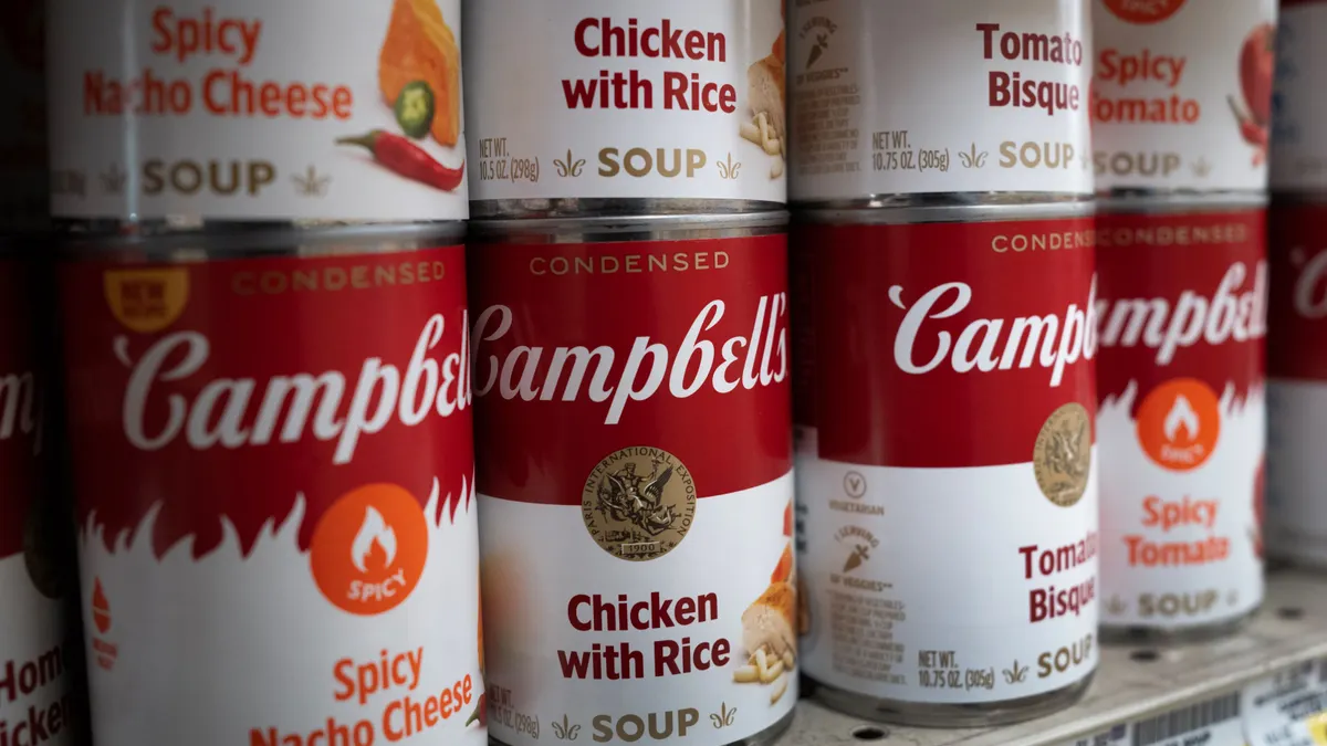 Four stacks of cans of Campbell's soup displayed on a grocery store shelf on June 2, 2025, in Chicago. Cans in the stacks, from left to right, are labeled "Spicy Nacho Cheese," "Chicken with Rice," "Tomato Bisque" and "Spicy Tomato."