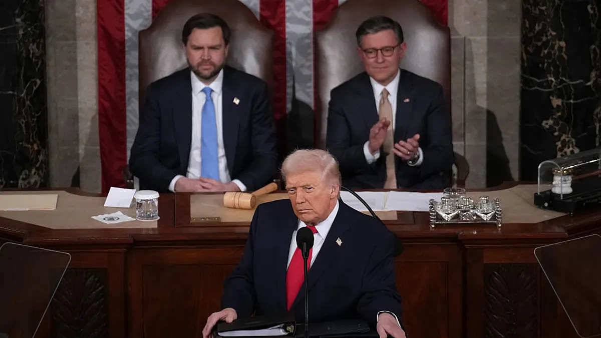 President Trump, wearing a red tie and a glower, is flanked by VP JD Vance and House Speaker Mike Johnson behind a podium.
