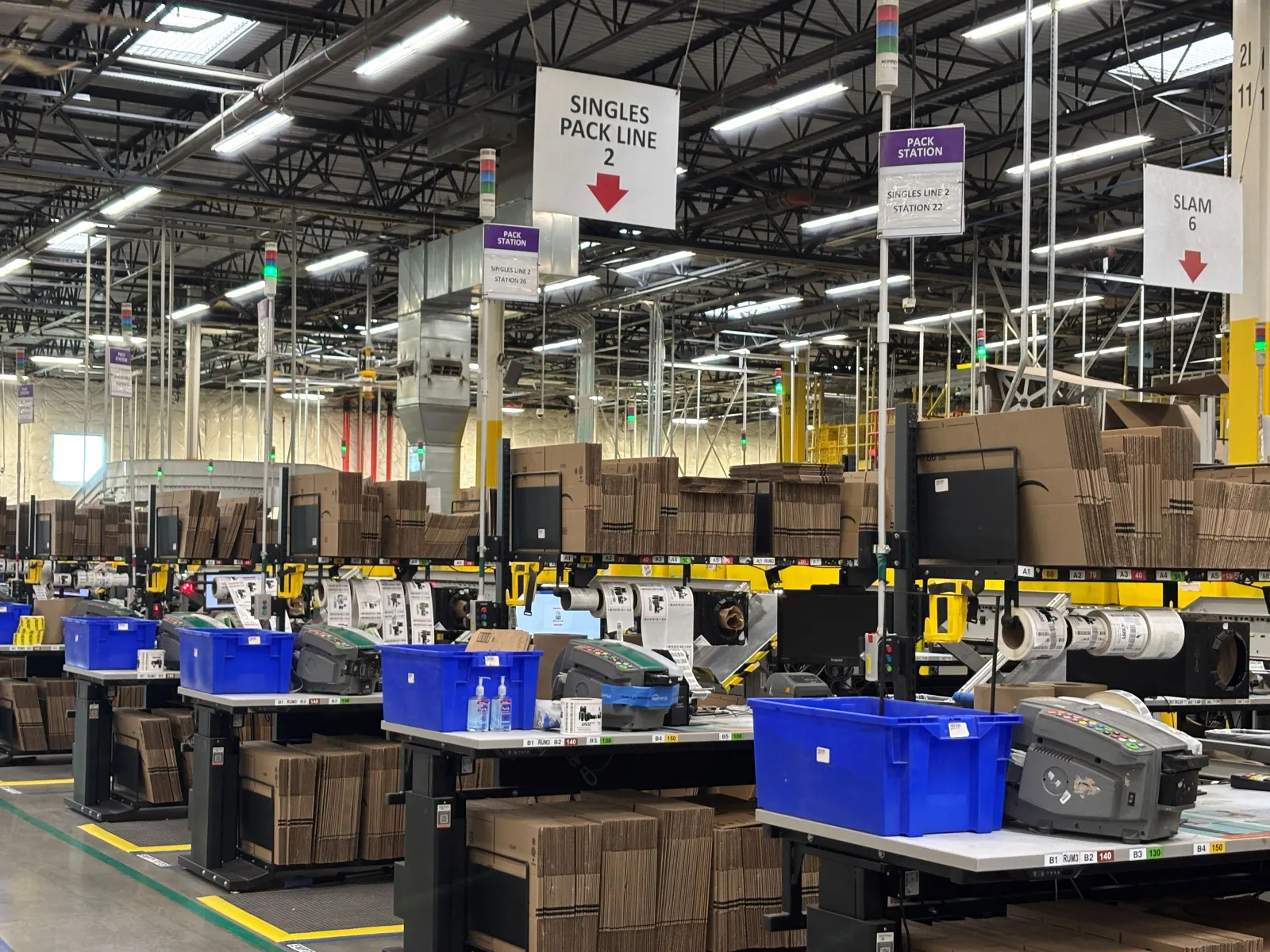 Folded cardboard boxes sit at workstations alongside blue bins in a warehouse.