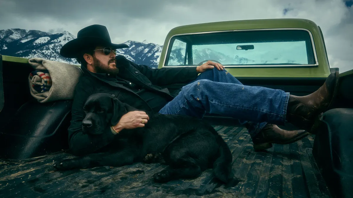 A man reclines in the back of a pickup truck wearing a cowboy hat, jeans and cowboy boots