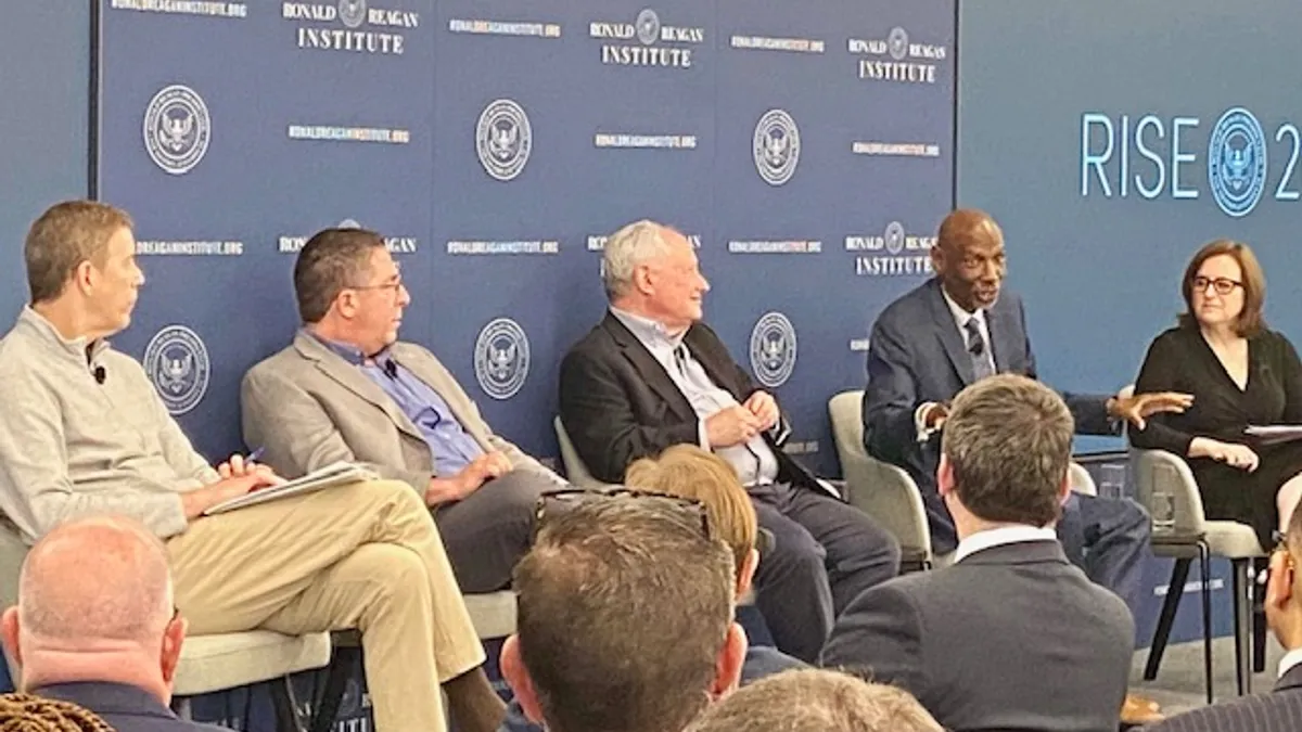 Five adults are sitting in a row on a stage in front of an audience. One person is speaking and the other adults are looking at the speaker.