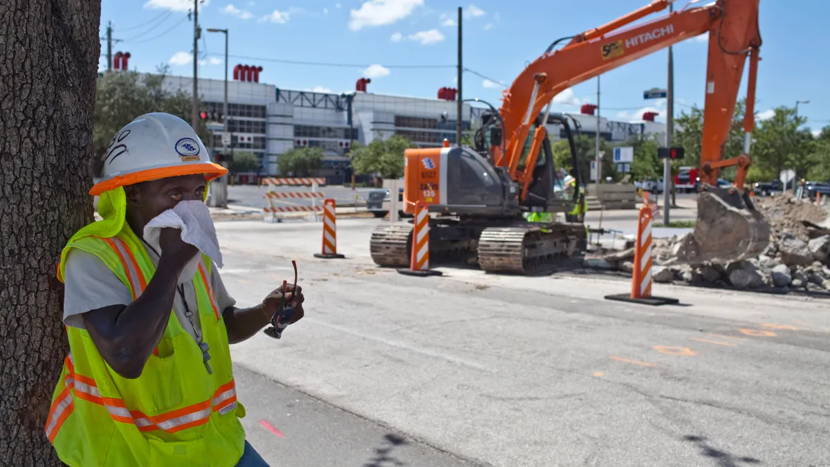 A construction worker in a reflective vest wipes his face as he shelters under a tree, while construction machinery and other workers stand in the background.