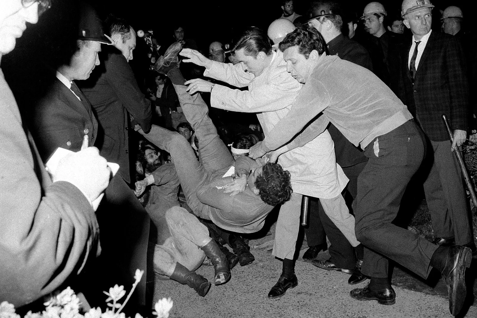 A protester falls back-first toward the ground while surrounded by police officers.