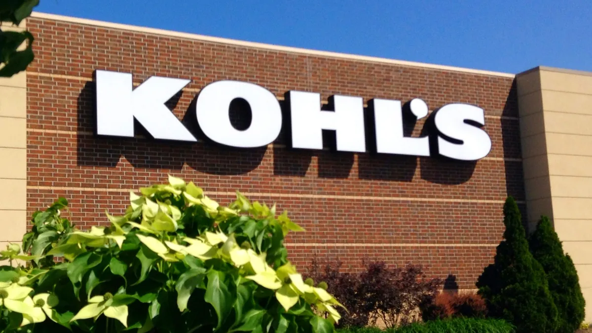 A leafy green bush sits in front of a reddish brick store against a bright blue sky, with the name Kohl's in white block letters.