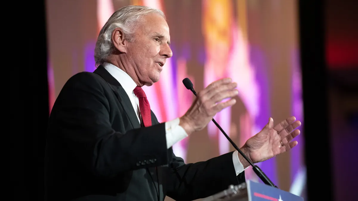 A side profile of South Carolina Gov. Henry McMaster standing at a podium with his hands raised and a partial view of a large screen behind him.