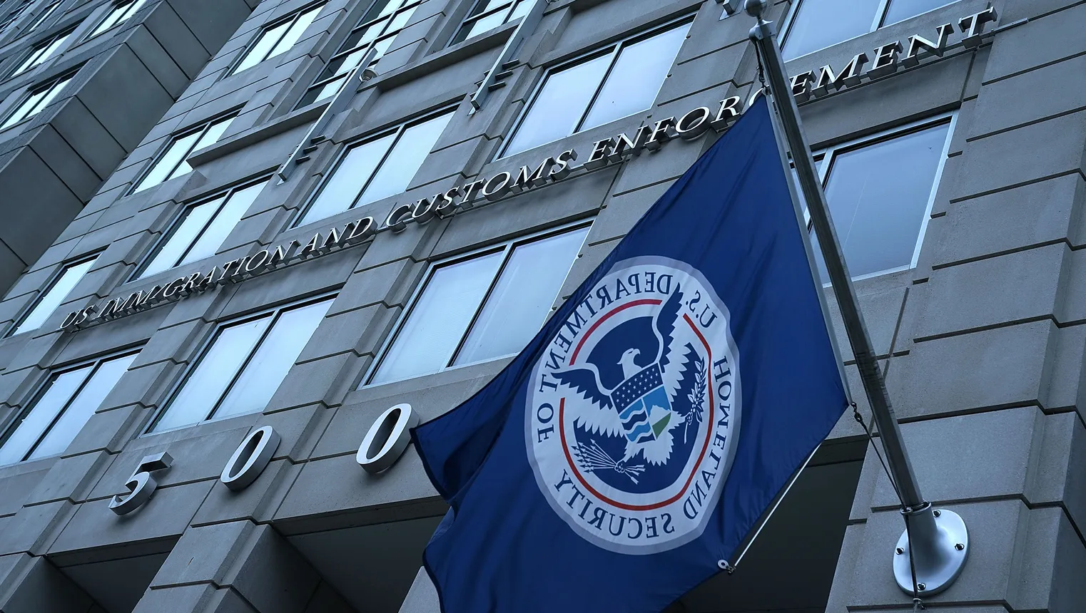 The exterior of the building is seen with a foreground DHS flag.