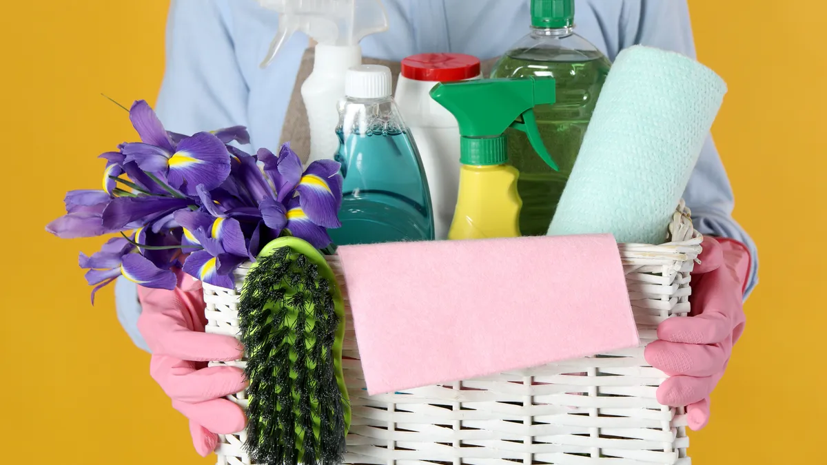 Person holding basket with detergents, flowers and tools on orange background, closeup