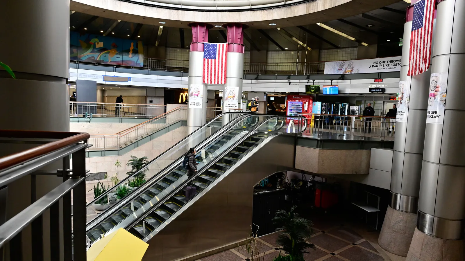 A person ascends one of two escalators with round silver pillars, an American flag and people on the upper level.
