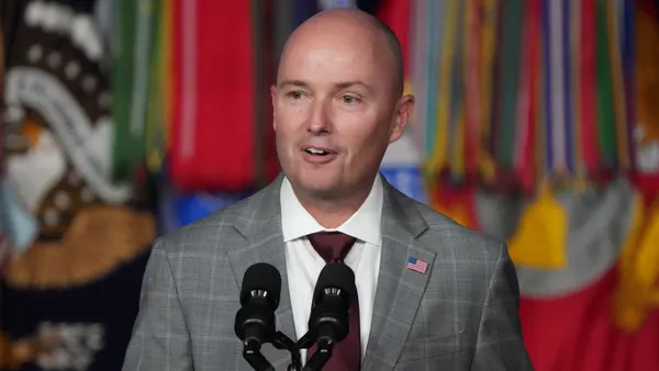 A person is standing at a podium with a microphone. Colorful flags can be seen int he background.