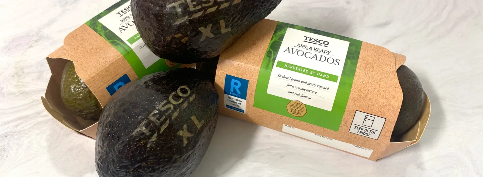 Two avocados with grocery brand Tesco&#x27;s messaging laser engraved onto their skin, and other avocados in fiber packaging.