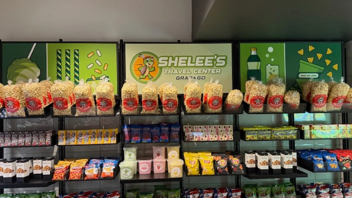 A photo of a rack of snacks inside a store, including popcorn, chips and candy. A sign above the snacks says "Shelee's Travel Center grab and go."