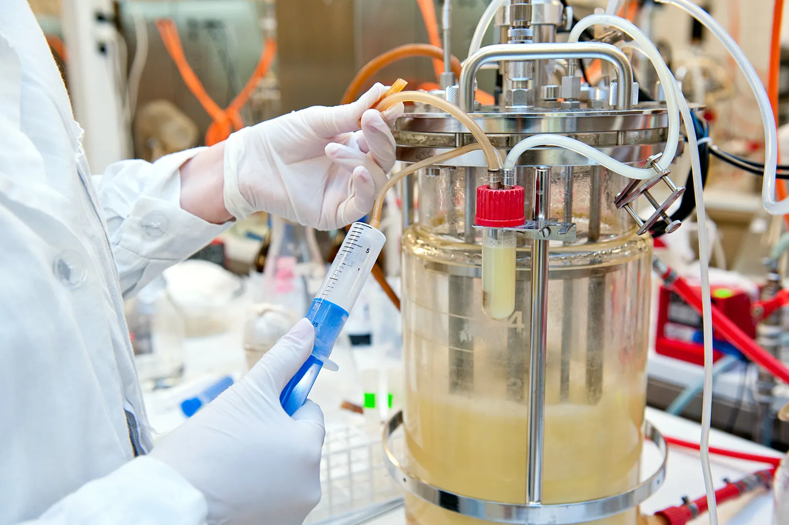 A person in a lab coat and gloves takes a sample of yellowish liquid from a small glass bioreactor in a lab setting.