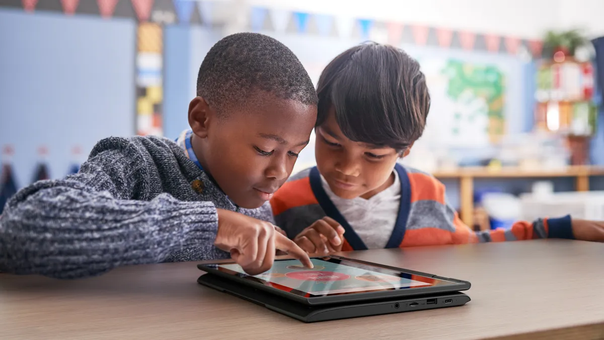 Two kids in a classroom playing on a tablet at a desk