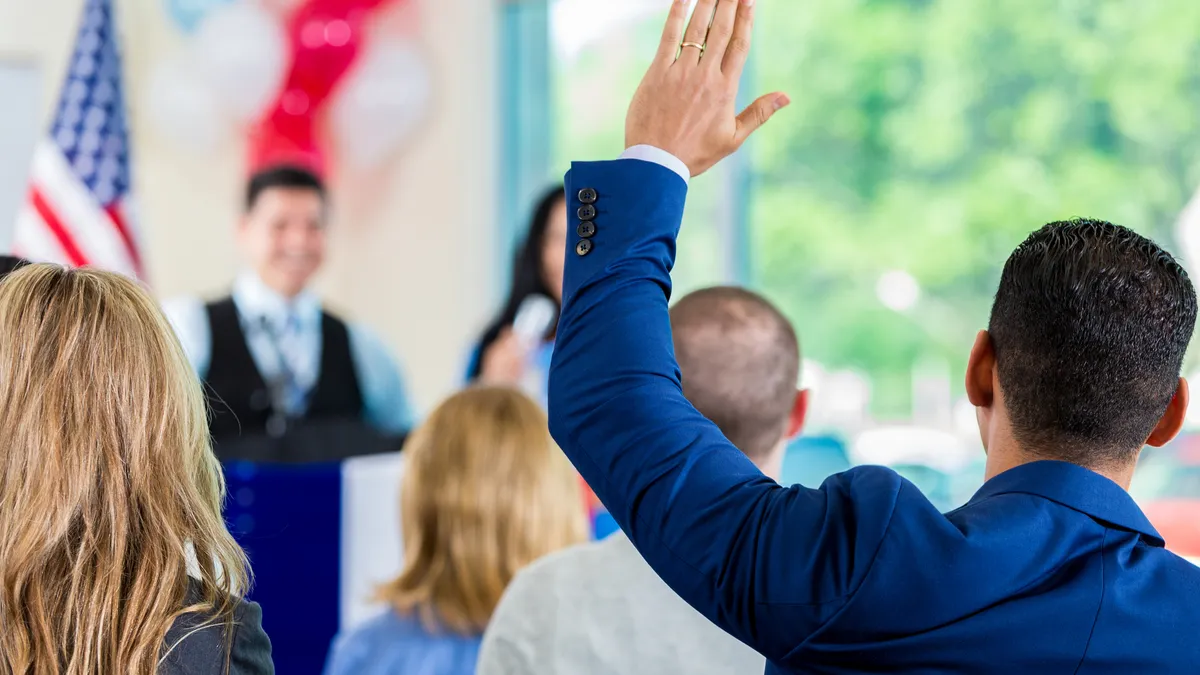 Mid adult Hispanic man is raising his hand to ask a question, while sitting in audience during political rally or town hall meeting. Speaker is standing at podium addressing crowd.