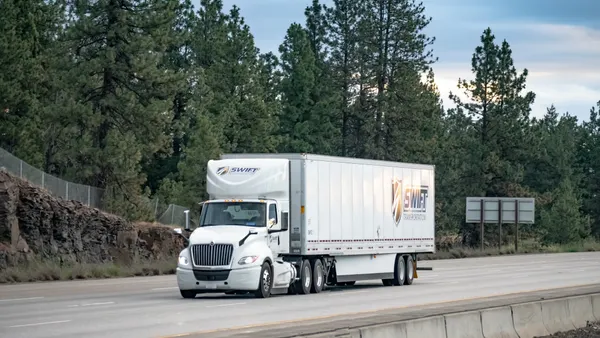 A  tractor-trailer is the sole vehicle shown on an interstate with trees in the background.