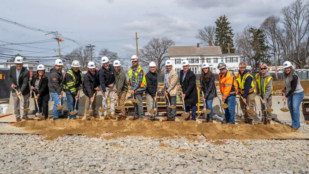 People with hard hats stand with shovels in a pile of dirt.