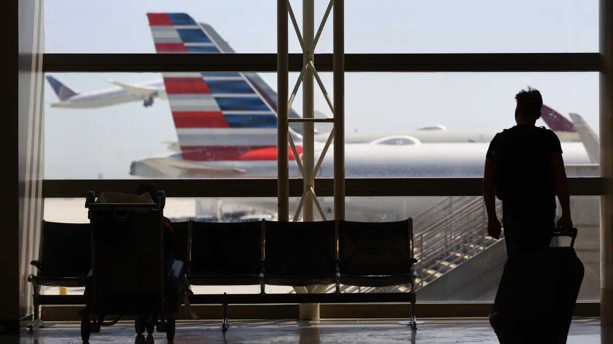 A person walks with luggage in front of window. Through the window, a plane sits at the gate and another is in the air.