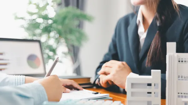 Two people in business attire sit next to models of apartment buildings and paperwork.