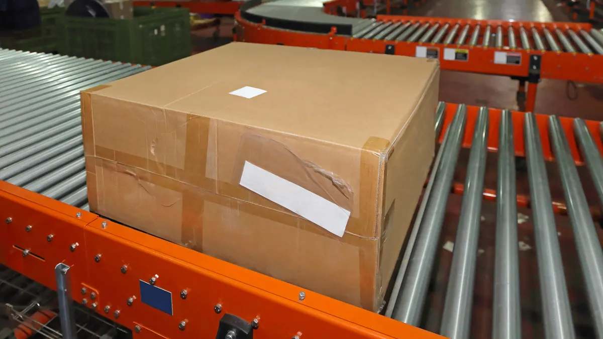 A sealed brown cardboard box with a white shipping label and packing tape sits on a red and silver metal conveyor belt system in a warehouse. Background includes industrial shelving, other packages, and factory equipment. The floor is polished red and the setting is brightly lit.