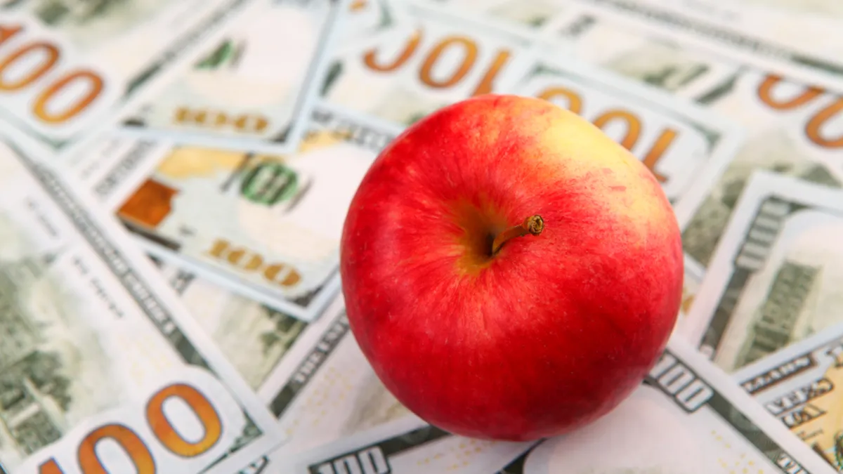 A red apple sits on top of a spread-out pile of $100 bills.