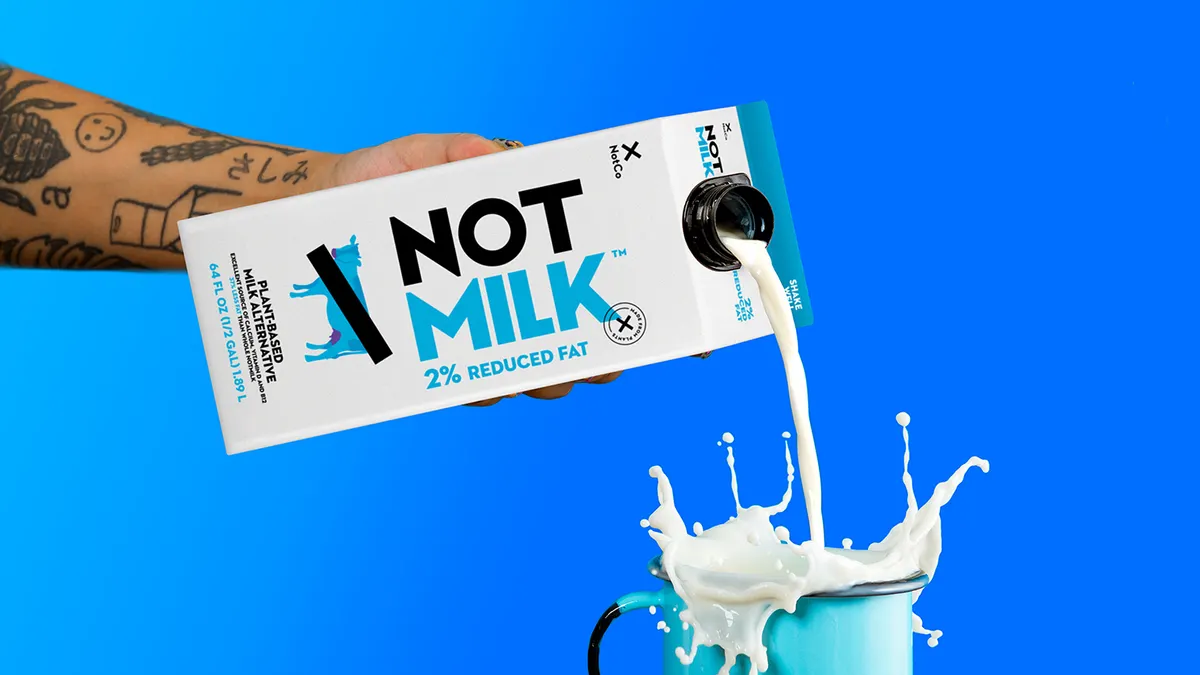 A hand pours 2% NotMilk from a carton into an overflowing blue mug.