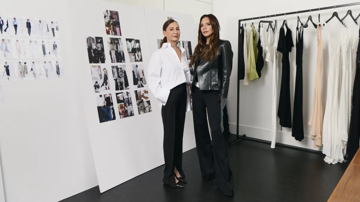 Two people stand next to each other in a studio in front of a rack of clothing.