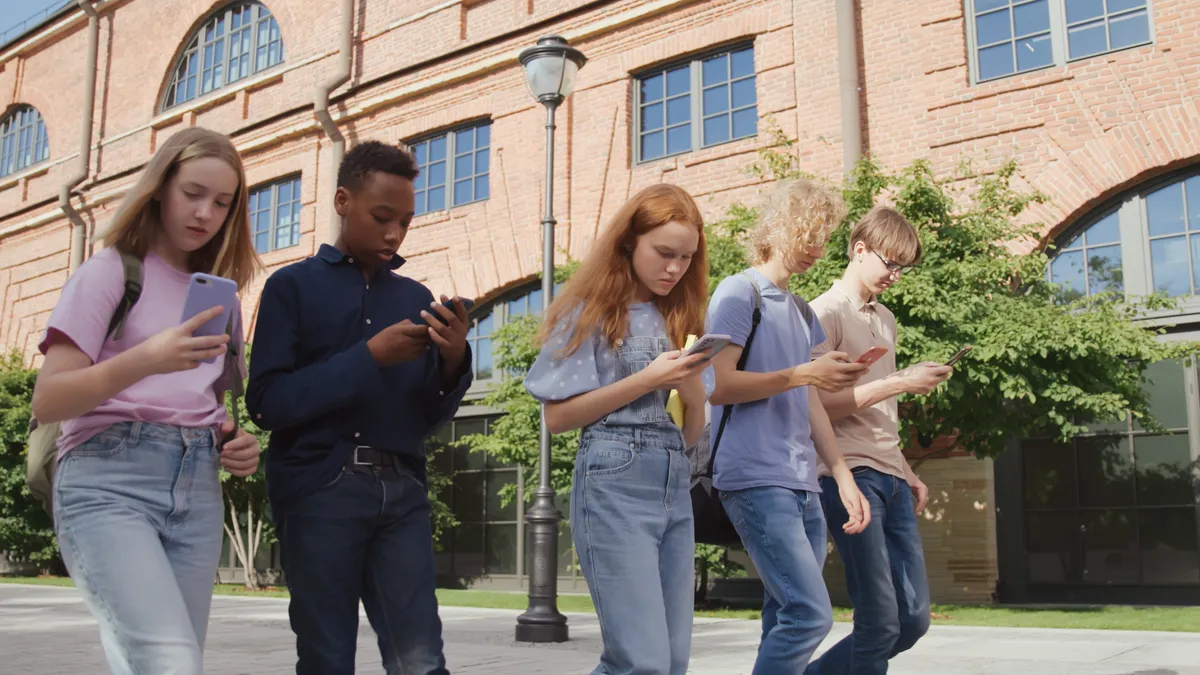 A handful of students walk in a row outside near a school building. They are each holding a cellphone and looking down at the cellphone.