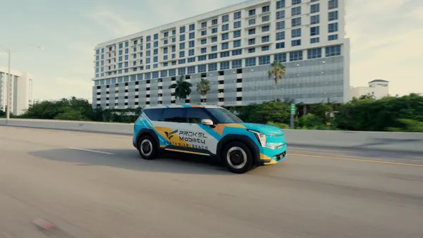 A green, blue and white SUV lettered "Prokel Mobility" on a road in front of a multistory building.