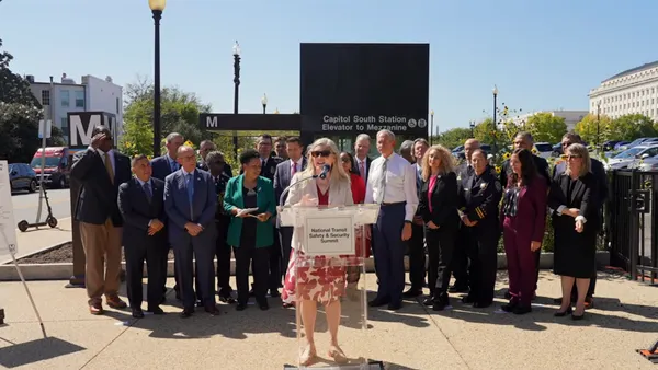 A person at a podium outdoors with a large group of people behind.