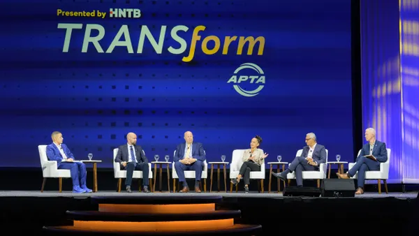 Six people on a stage seated in white charge under a sign reading "TransForm."