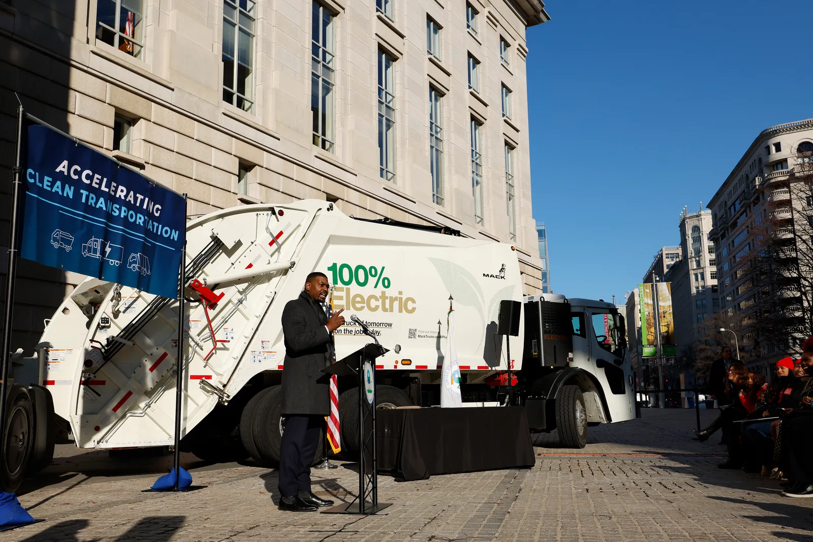 Man in jacket stands in front of electric white garbage truck