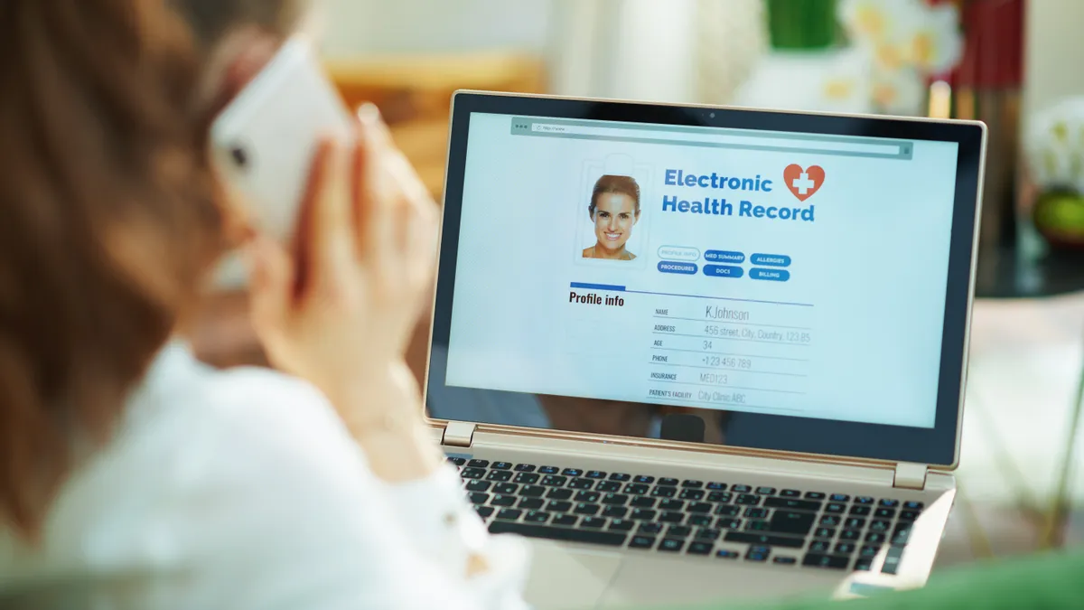 Seen from behind, a clinician is using a cell phone and checking a patient's electronic health record on a laptop.