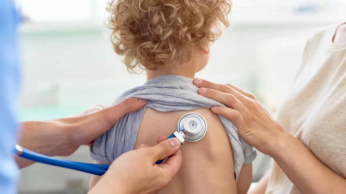 A child has their back to the camera and their shirt is pulled up to the next. On either side of the child are adult hands. One adult is holding a stethoscope to the child's back.