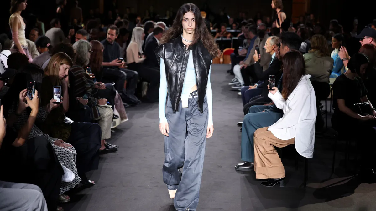 A model walks the runway while onlookers are seated on either side. The model wears baggy pants and a leather vest.