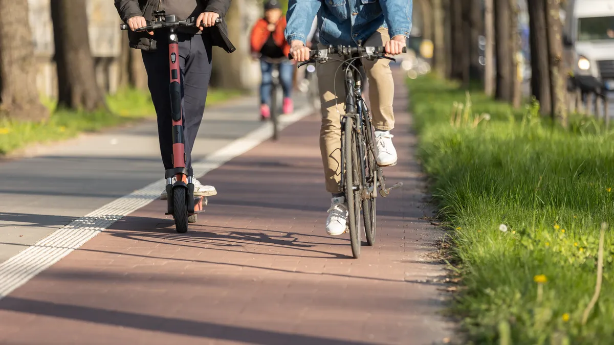 Two people are seen riding a scooter and a bicycle along a path.