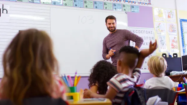 A teacher stands in front of a classroom white board with the number "103" written. A row of young elementary school students sit and listen in front of the teacher.