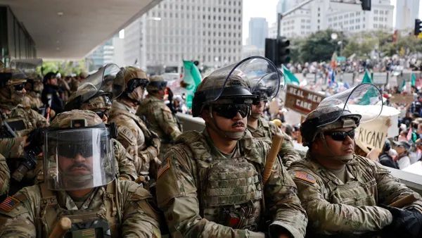 People wearing military uniforms and helmets with a crowd of protesters behind them.