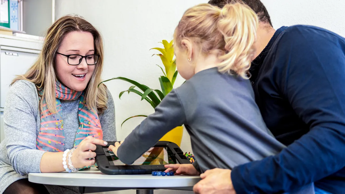 An adult is seated at a desk. On the opposite side of the desk sits a young child on the lap of an adult. The child is touching a device on the desk.