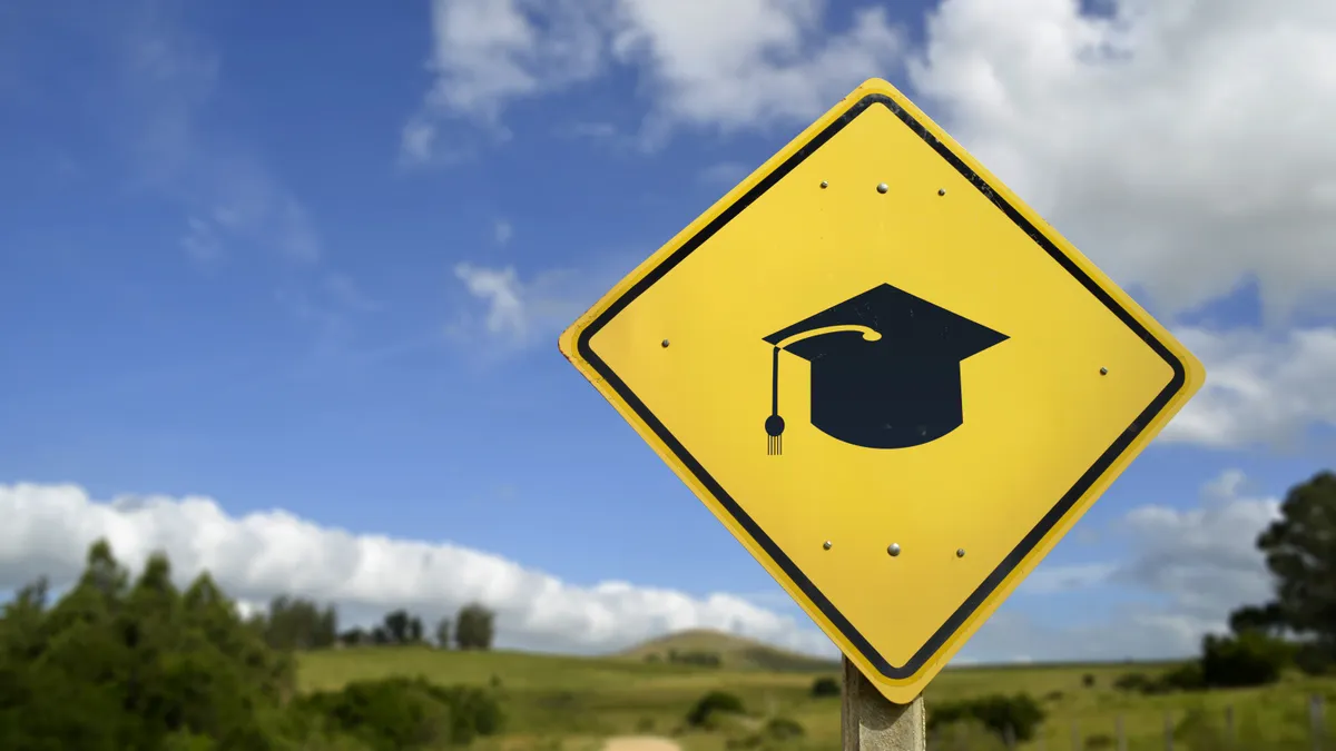 A road sign with a graduation cap icon stands in a rural, grassy landscape.