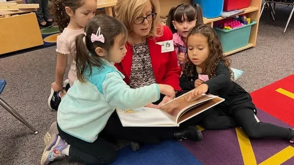 An adult sits on a carpeted floor in a classroom. They are surrounded by  young children who are looking at a large picture book in the adult's lap.