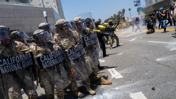 A line of people wearing camouflage with large shields that say "California National Guard" face a crowd.