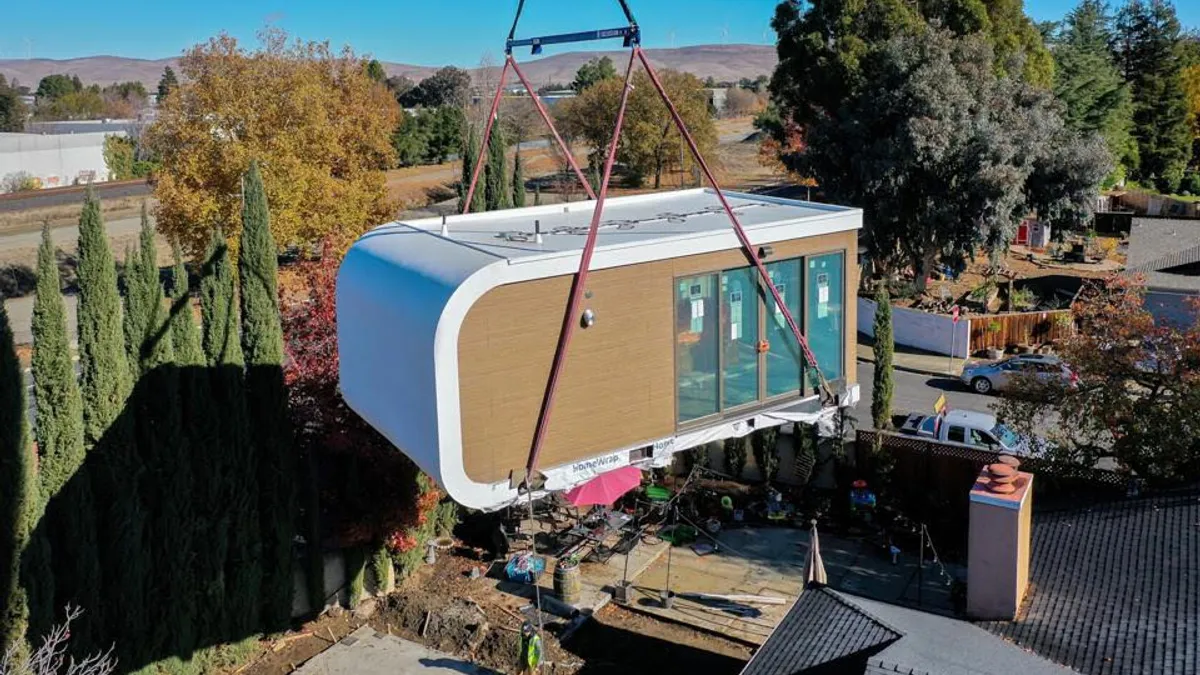 A 3D-printed home module is being slowly let down by an industrial machine that has straps around the building. It's a sunny day in a residential area.