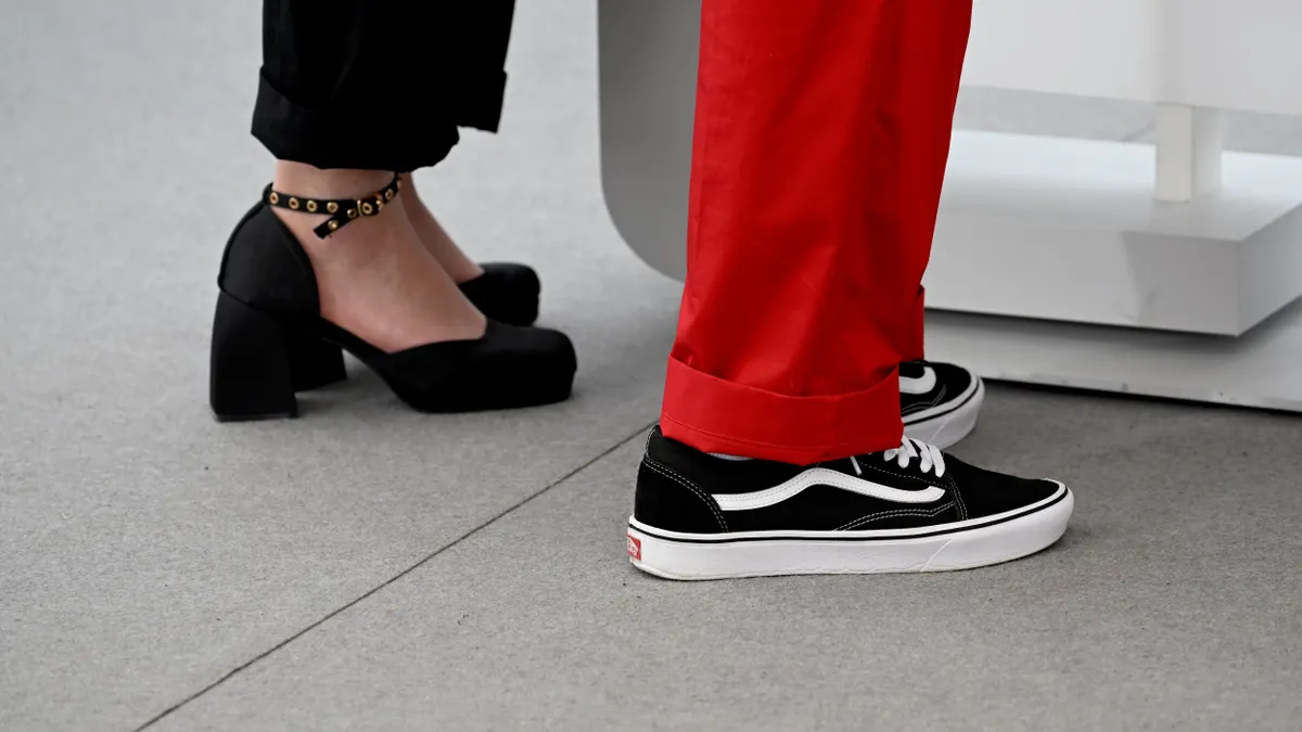 A close up of two people's shoes, one wearing heels, then other Vans.